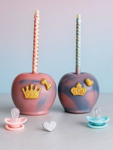 Zwei gefärbte Äpfel mit Kronen- und Herzmotiven sowie Schnullern in Pink und Blau.